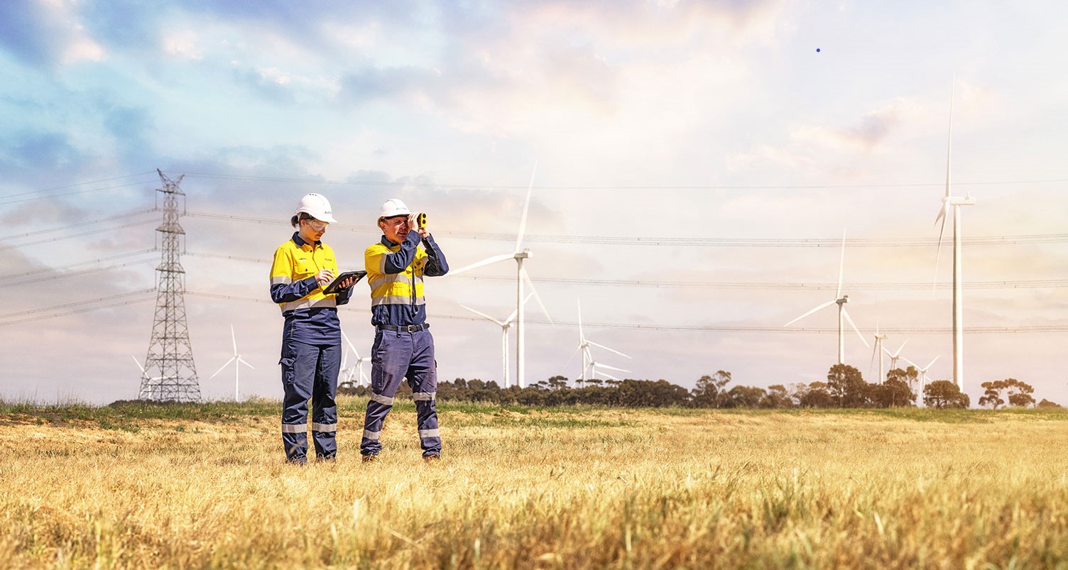 Two workers standing in a field near electricity transmission towers and windmills